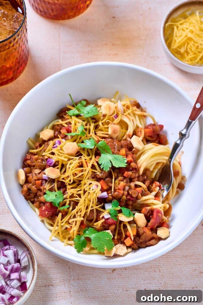 Cincinnati-Style Lentil Chili Recipe for Rich, Hearty Flavor 2 A bowl of lentil's chili over spaghetti and garnish with oyster crackers, red onion, cilantro and shredded cheese.