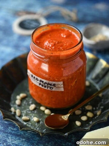 Red pepper pesto in a jar with grated cheese in the front on a blue surface.