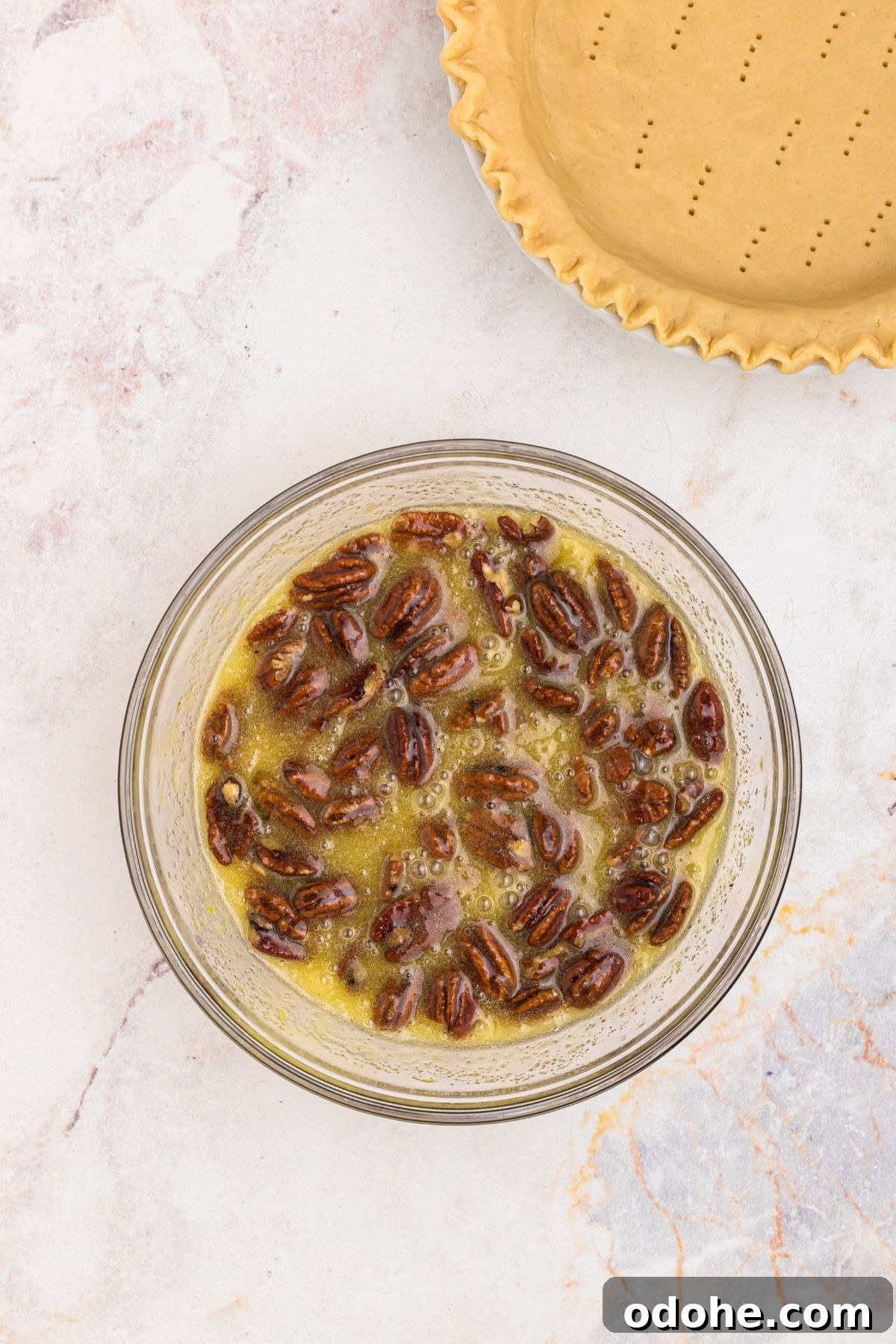 Clear glass bowl with egg and corn syrup mixture and pecans mixed together, next to a pie crust.