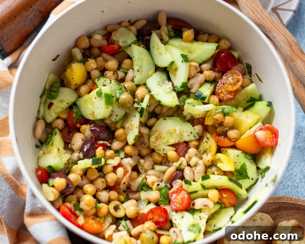 horizontal photo of a white serving dish with a bean salad made with cannellini beans and chickpeas with olives and summer vegetables like cherry tomatoes, cucumber, celery, and fresh basil.