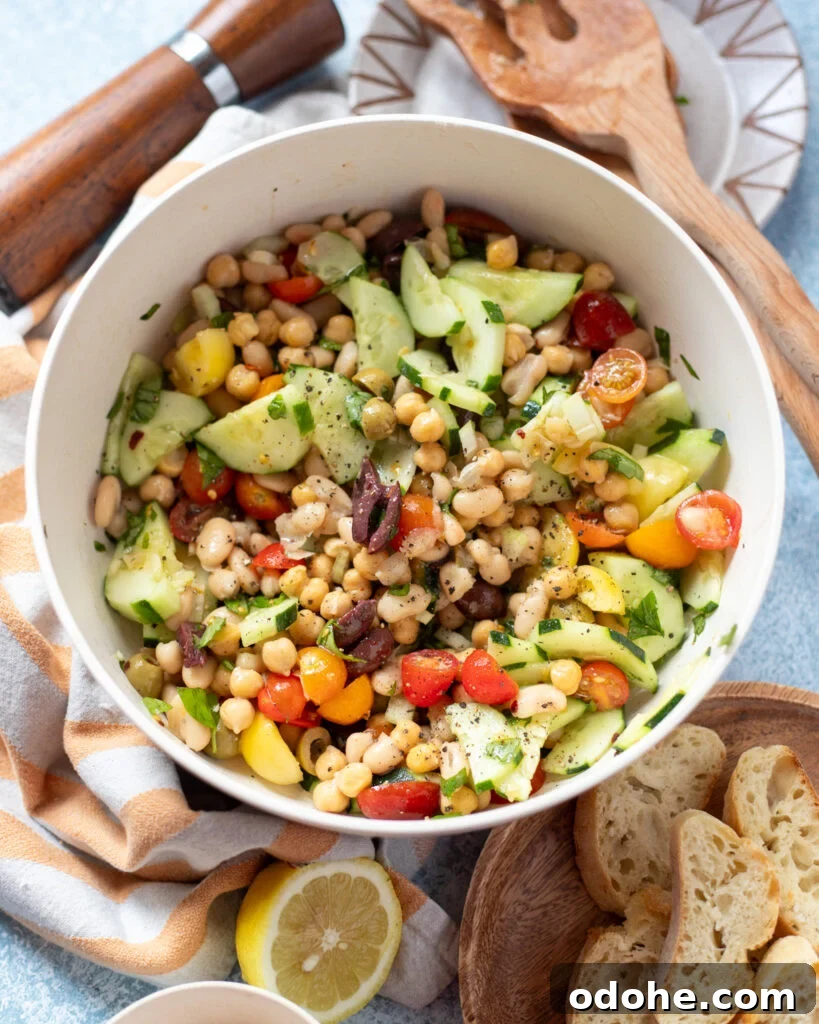 a white serving dish holding a vegan dense bean salad with Mediterranean seasonings and vegetables next to sliced French bread and serving utensils.