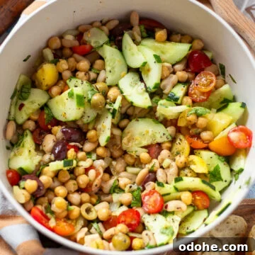overhead view of a white serving bowl holding a dense bean salad made with two types of canned beans, cucumber, tomato, celery and olives in a simple lemon and olive oil dressing.