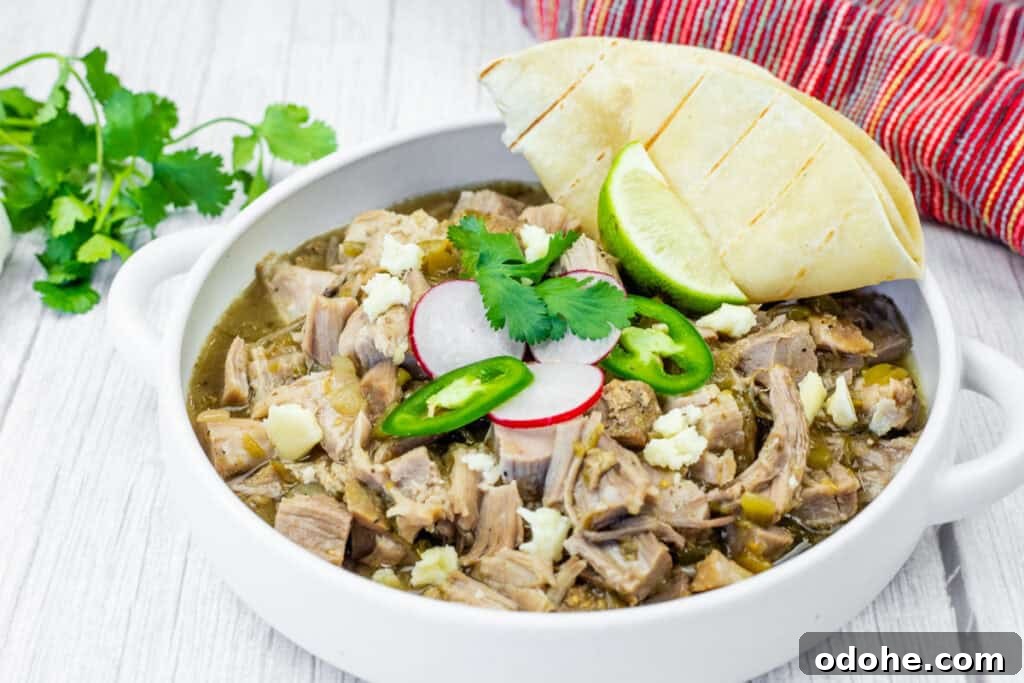 Slow Cooker Pork Chile Verde Recipe for Tender, Flavorful Pork 8 A white bowl of Slow Cooker Pork Chile Verde, featuring visible chunks of meat, radish slices, jalapeno slices, cilantro, and cheese, sits on a white wooden table. A folded tortilla and lime wedge are placed beside the bowl. A head of garlic and cilantro sprigs are in the background, along with a red and white striped cloth.