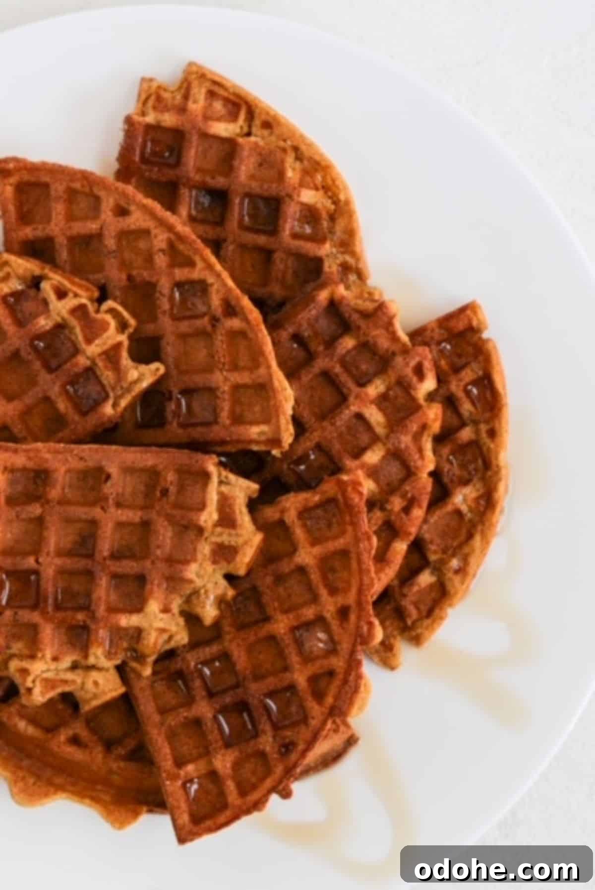 Several quarters of round pumpkin waffles overlapping on a white plate and topped with a drizzle of maple syrup.