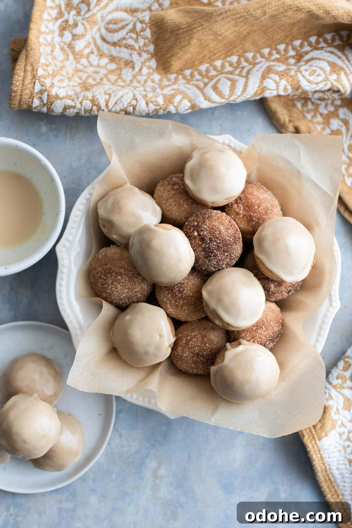 A mix of glazed and sugared donut holes on a bowl next to glaze.
