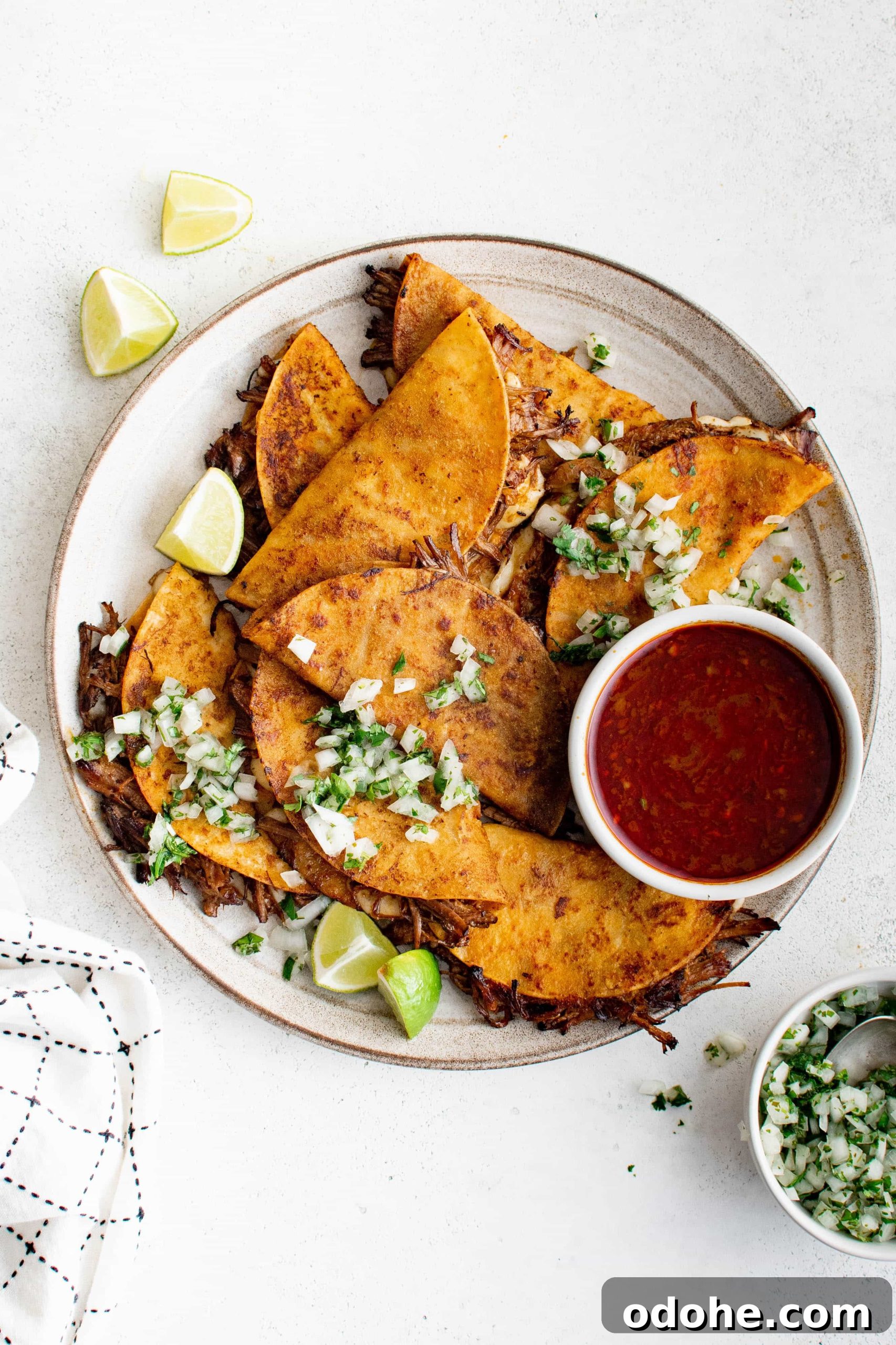 40+ Authentic Mexican Recipes for an Unforgettable Cinco de Mayo 13 White plate filled with birria tacos garnished with onions ,cilantro, and lime wedges, and served with a small bowl of birria broth consumé.
