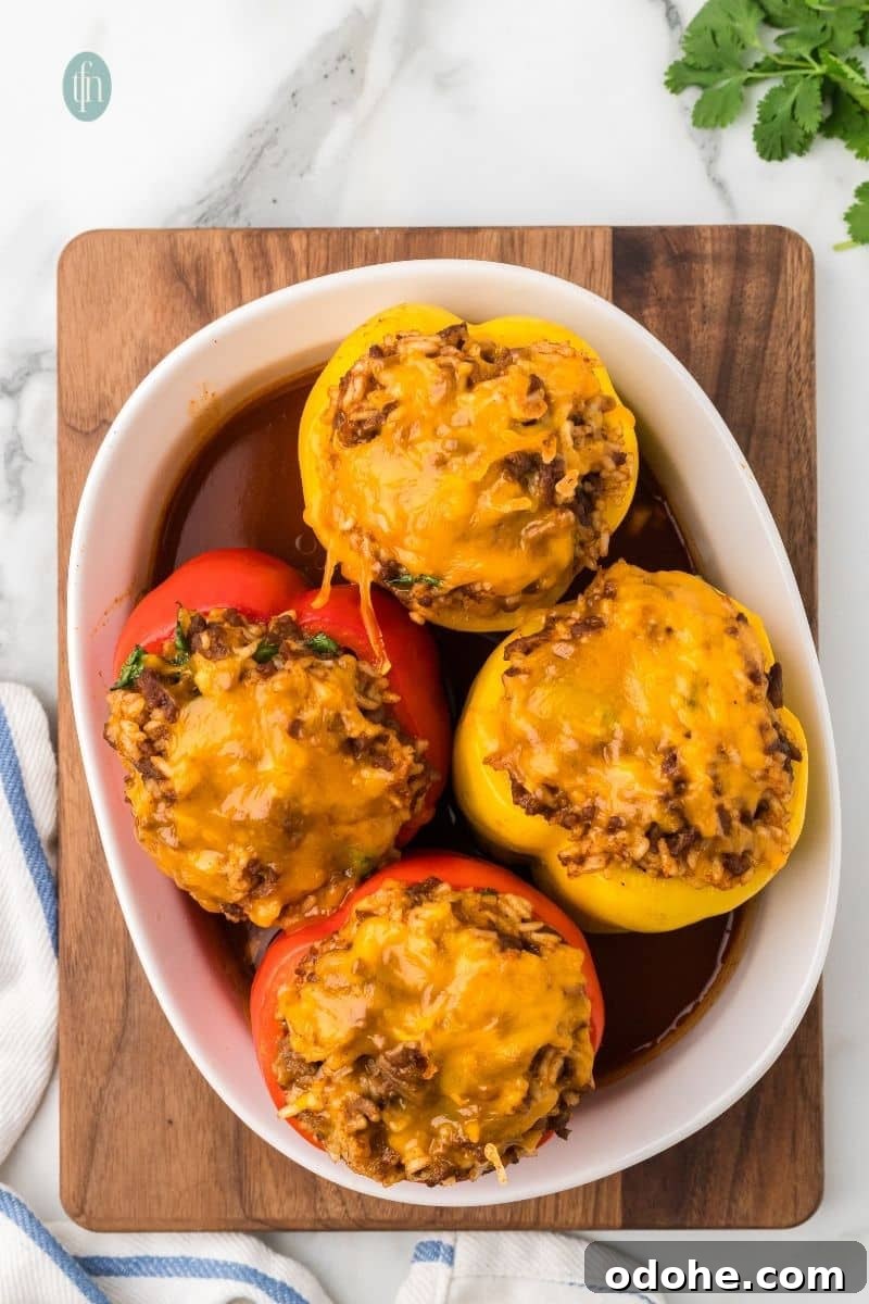 An overhead shot of four baked stuffed bell peppers in a white oval baking dish, sitting on a wooden cutting board with a blue and white striped towel. The peppers are topped with melted cheese and sit in a pool of sauce.