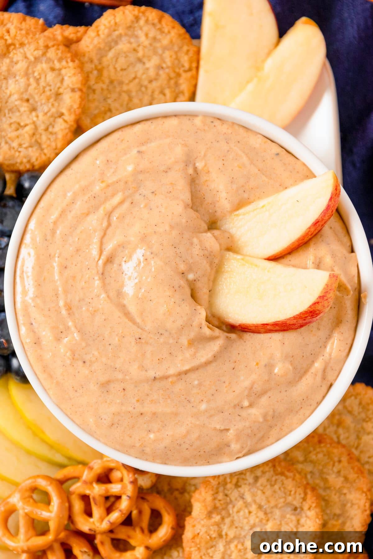 Pumpkin dip in a white bowl topped with a sliced apple fan and served with graham crackers and pretzels.